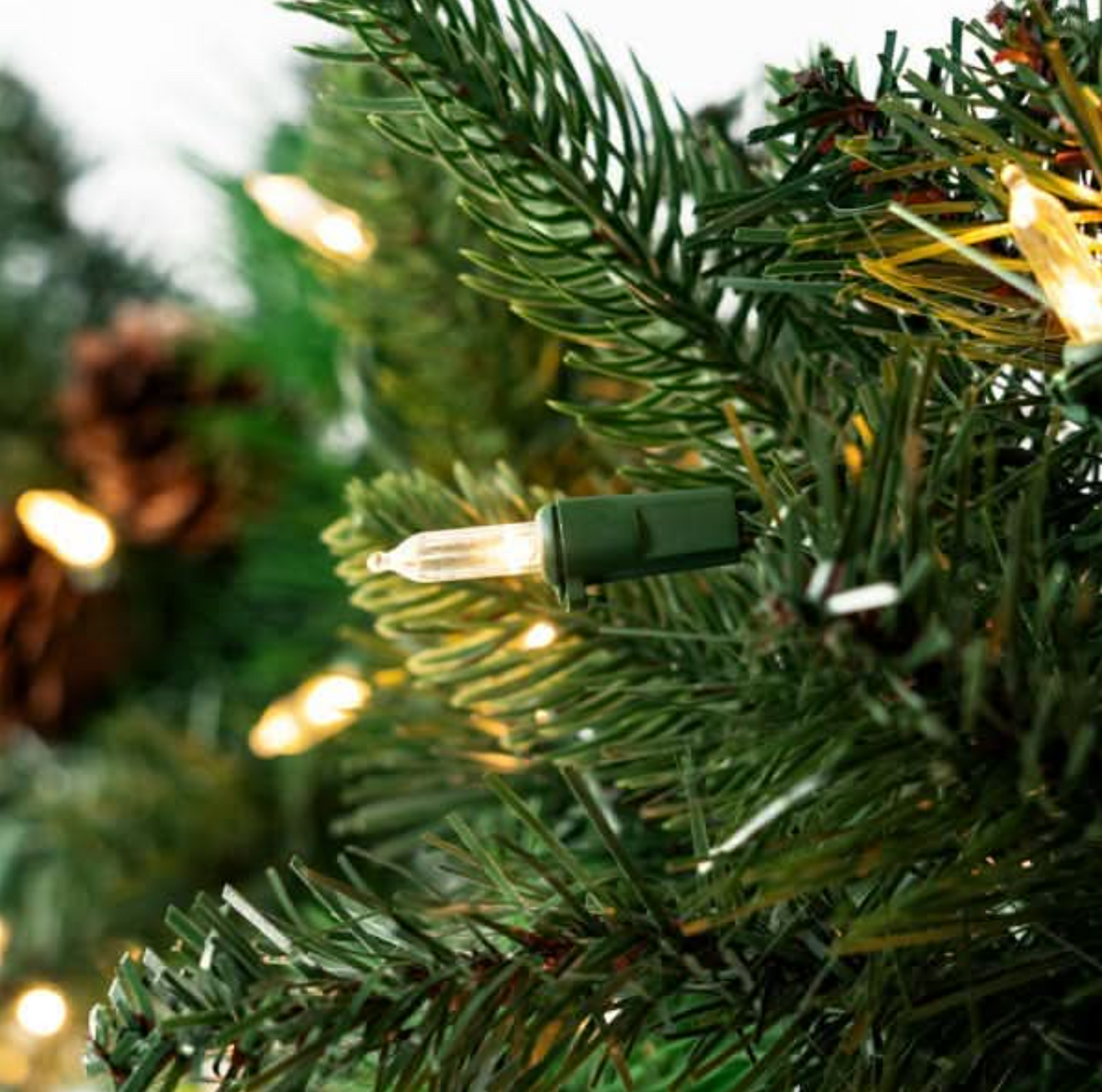 Close-up of warm white Christmas tree lights wrapped around green artificial pine branches, with soft bokeh lights in the background.