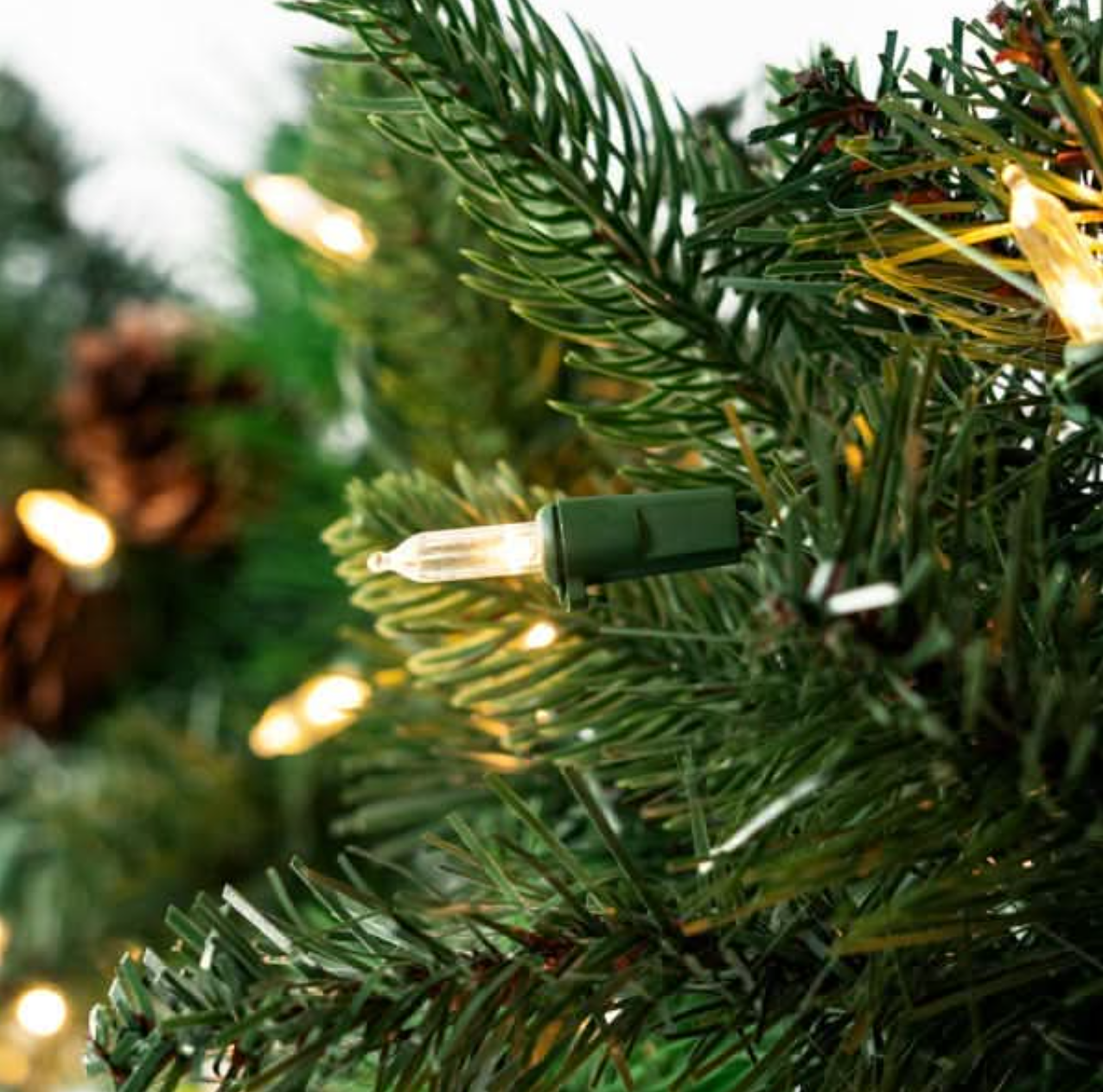 Close-up of warm white Christmas tree lights wrapped around green artificial pine branches, with soft bokeh lights in the background.